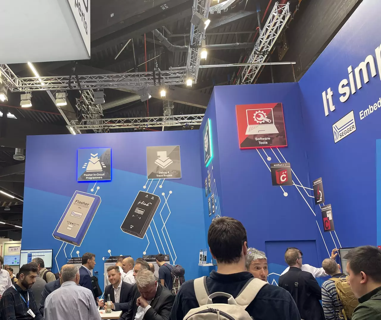 A busy exhibition booth featuring a blue backdrop with various product icons and information. Attendees engage in discussions around tables, showcasing technology tools. Notable items include circuit programming devices and software tools, indicating a focus on embedded systems and development solutions.