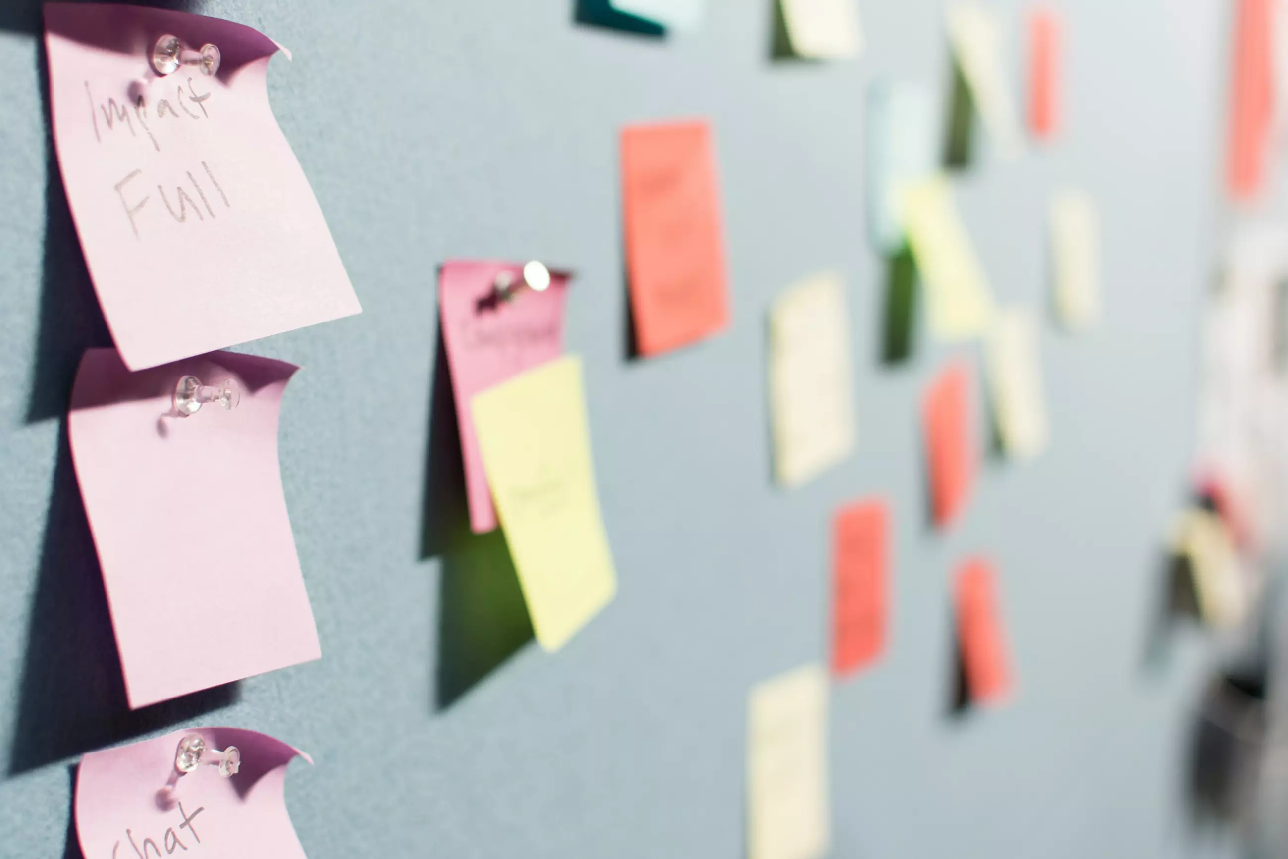 A bulletin board covered with colorful sticky notes in various colors, including pink, yellow, and green. Some notes are legible with words such as "Impact" and "Chat." The background is a blue wall, suggesting a brainstorming or planning session.