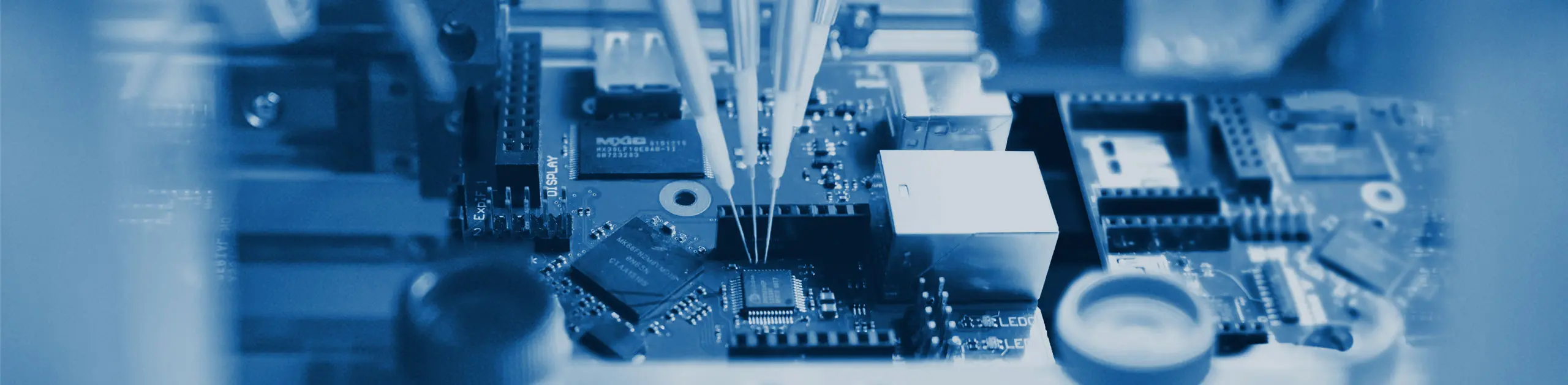 Close-up view of a circuit board featuring various electronic components, including chips and connectors. Two precision instruments are positioned over the board, indicating a process of assembly or repair in a technological setting.