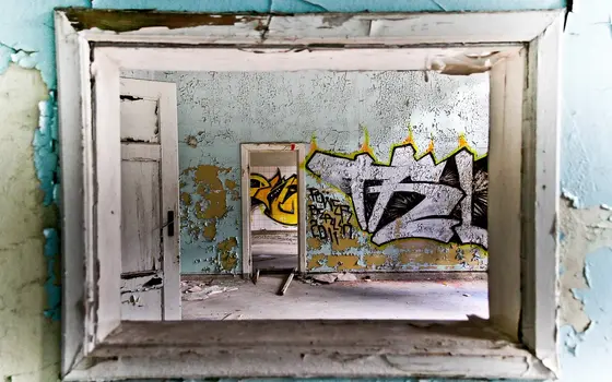 The image shows a view through a weathered window frame into an abandoned room. The peeling blue walls contrast with vibrant graffiti on the opposite wall, depicting abstract shapes and text. A second doorway is visible, leading deeper into the space.