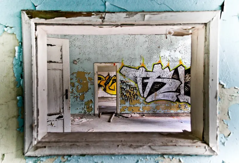 The image shows a view through a weathered window frame into an abandoned room. The peeling blue walls contrast with vibrant graffiti on the opposite wall, depicting abstract shapes and text. A second doorway is visible, leading deeper into the space.