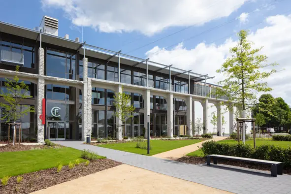 Arcade Building at Factory Campus The arcade building on the Factory Campus in Düsseldorf features large windows and an entrance area. In front of the building stretches a well-kept green area with small trees and a path made of gray paving stones. A bench is also visible. The sky is partly cloudy.