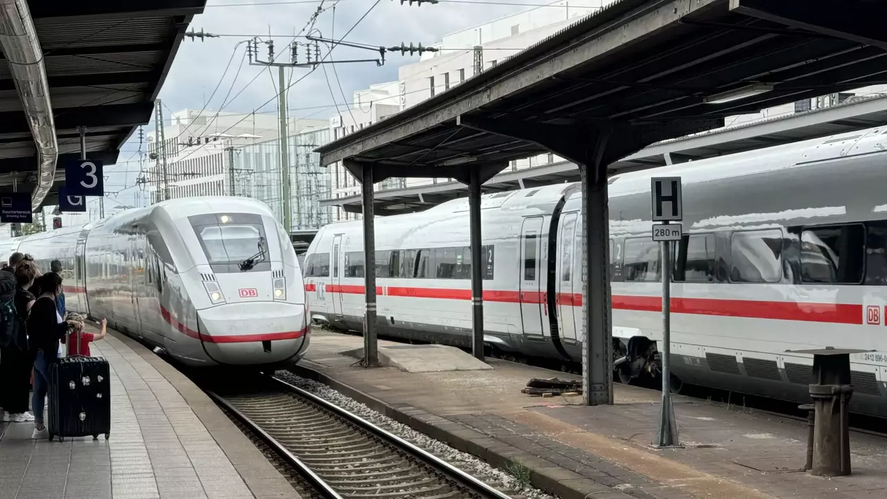 A modern high-speed train approaches a train station platform, with multiple carriages parked alongside. Passengers wait on the platform, which features tiled flooring and tracks visible leading away from the station. The scene conveys a busy transportation hub.