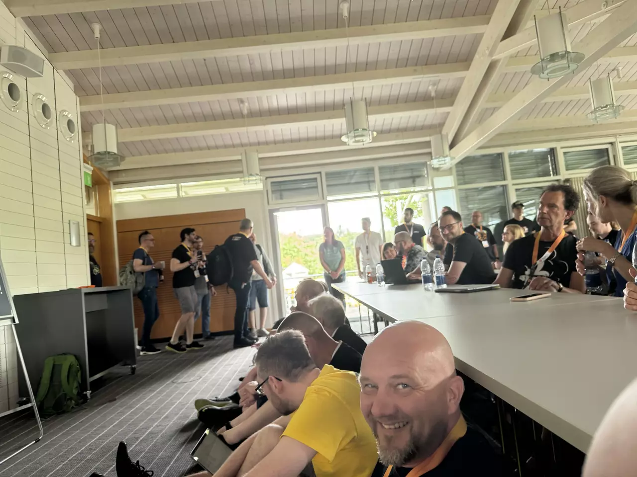 A group of people is seated in a spacious room with large windows. Some attendees are engaging in conversation, while others are entering from a side door. A smiling man in the foreground is wearing a yellow shirt. The atmosphere appears lively and social.