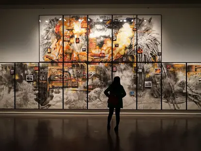 A person stands in front of a large, vibrant artwork consisting of multiple panels. The artwork features bold colors like orange and black, depicting abstract forms and smaller framed images scattered throughout. The scene captures the viewer's engagement with the artistic display.