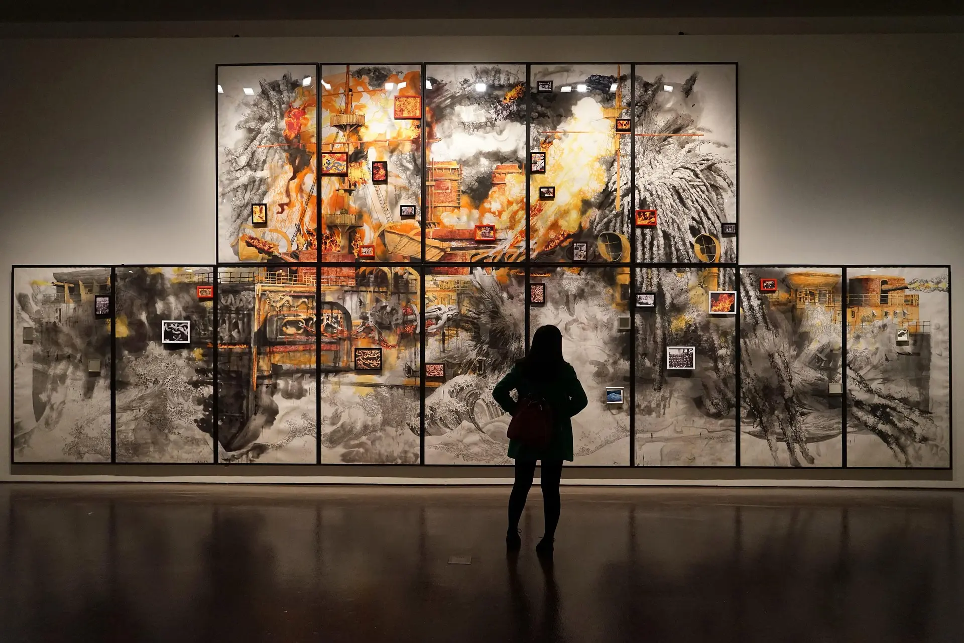 A person stands in front of a large, vibrant artwork consisting of multiple panels. The artwork features bold colors like orange and black, depicting abstract forms and smaller framed images scattered throughout. The scene captures the viewer's engagement with the artistic display.